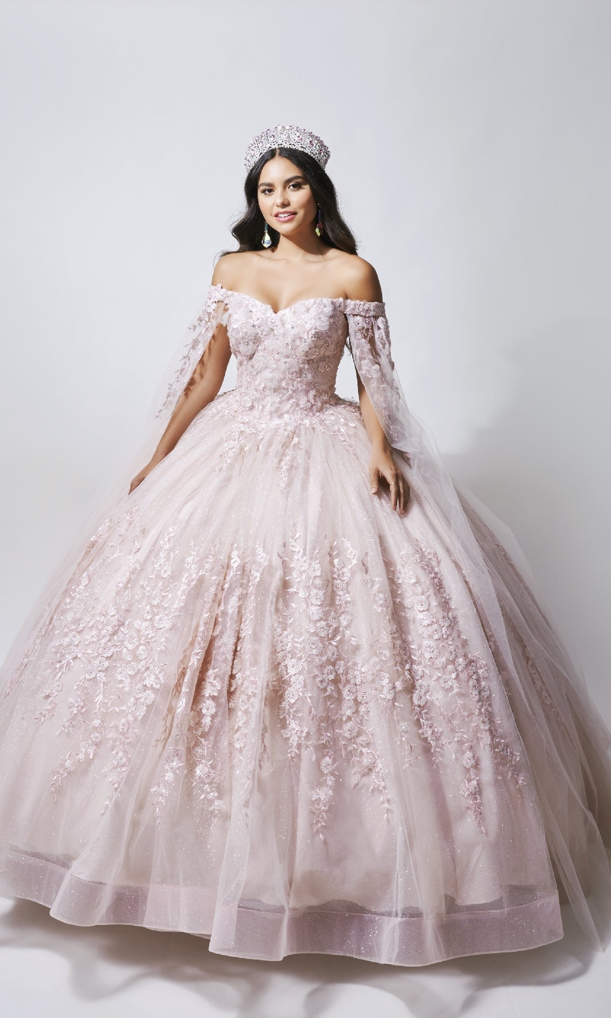 A woman in the Nina Canacci Q109 long quinceanera dress, an off-the-shoulder light pink gown with a floral bodice and matching crown, stands against a plain white background.